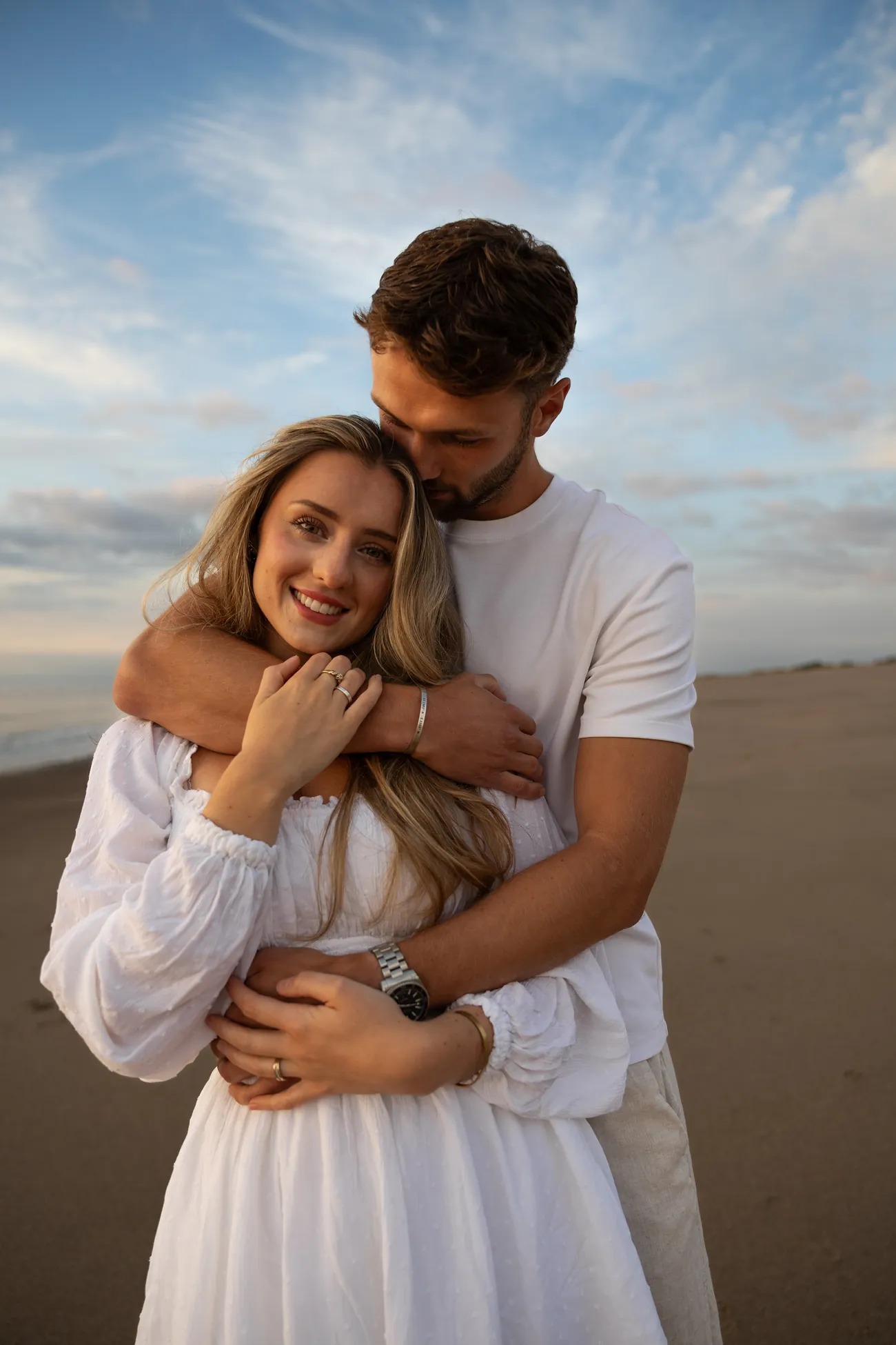 A man kisses his fiancée's forehead on a beach, with dunes and beach huts behind them