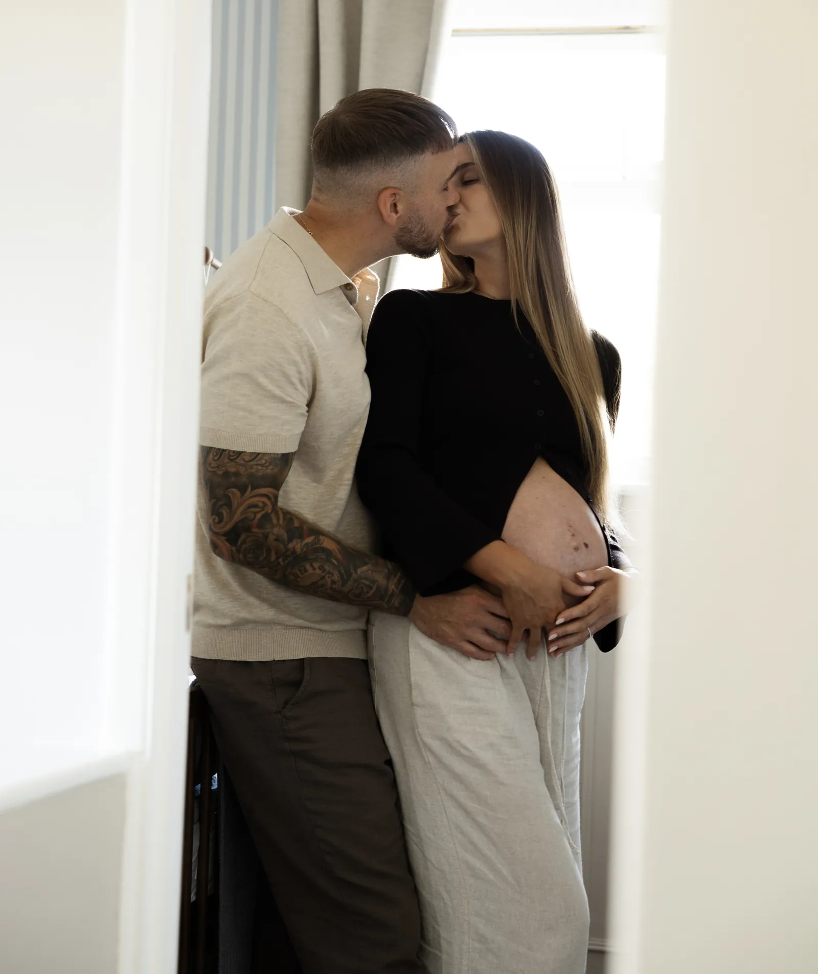 An expectant couple kiss by a sunlit window, her hands resting on her bump