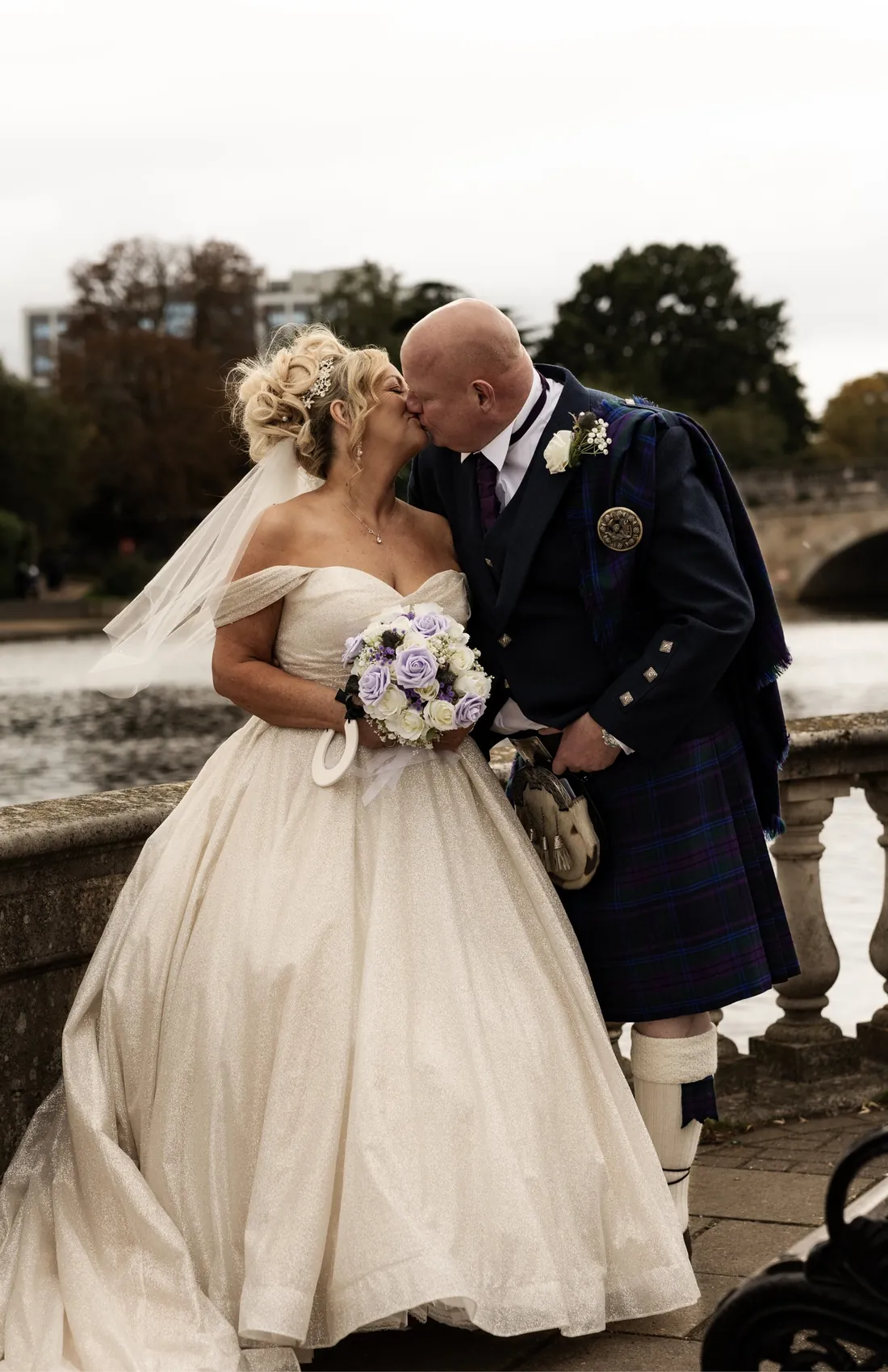 A bride and groom in a kilt kiss beside a lake, her ivory gown spread around her